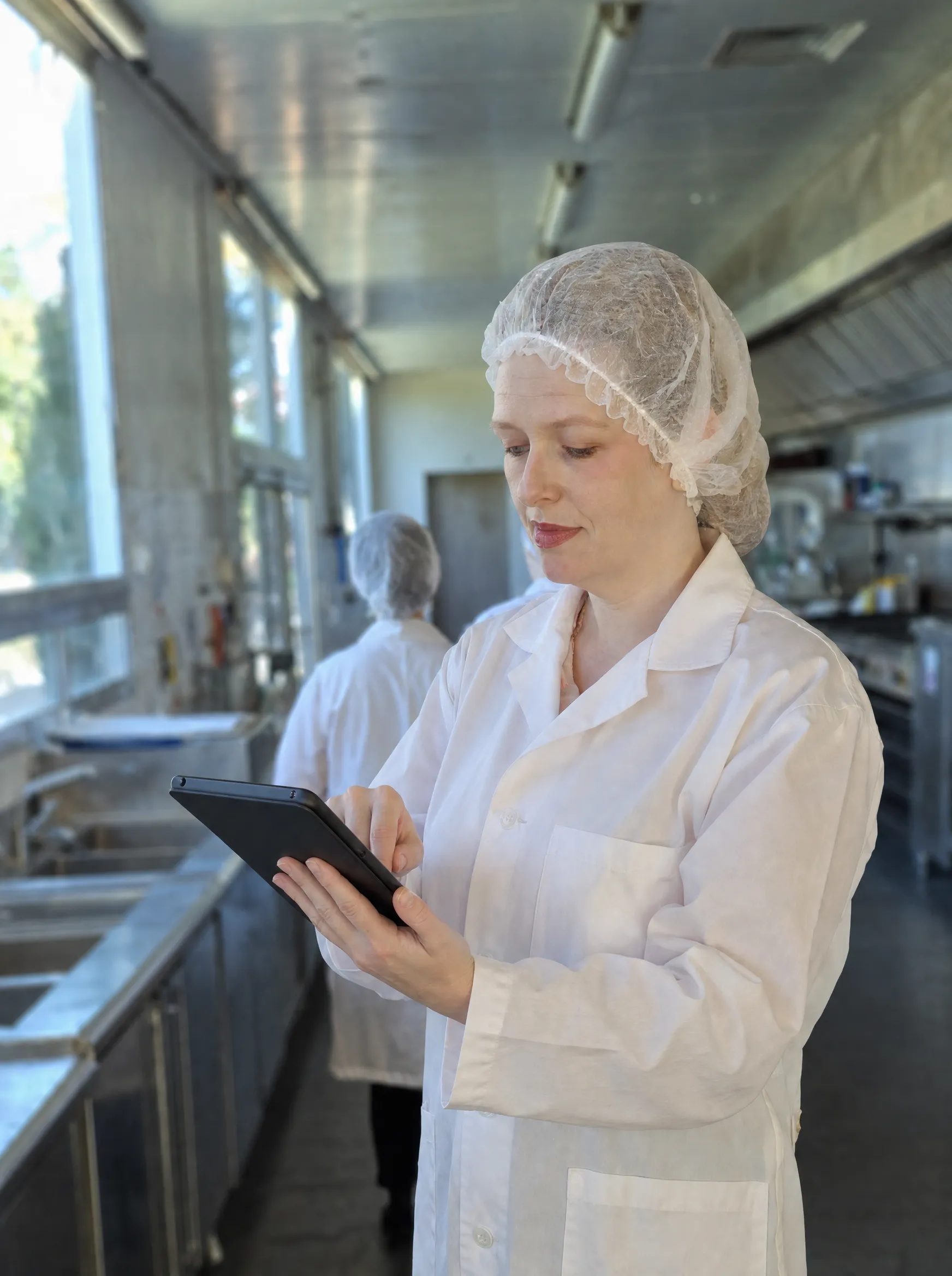 Suelen Defante inspecionando produção com tablet em cozinha industrial, EPI completo